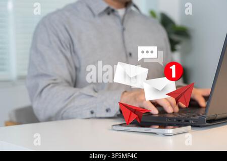 Person using a laptop with floating email, chat, notification icons above a smartphone, representing digital communication, email marketing, online me Stock Photo
