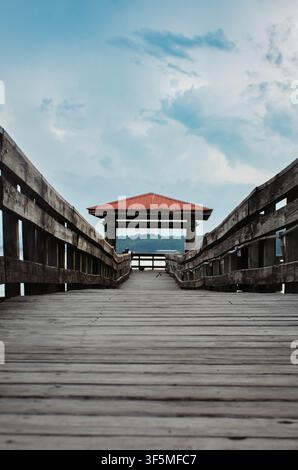 Empty wooden boardwalk pier leading to a covered pavilion over a calm lake under a cloudy sky Stock Photo