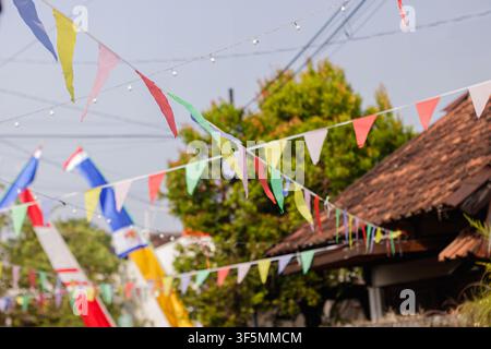 Close focus on multicolored triangular pennant flags hanging outdoors with a traditional village house roof and lush green trees in the background. High Quality Photo Stock Photo