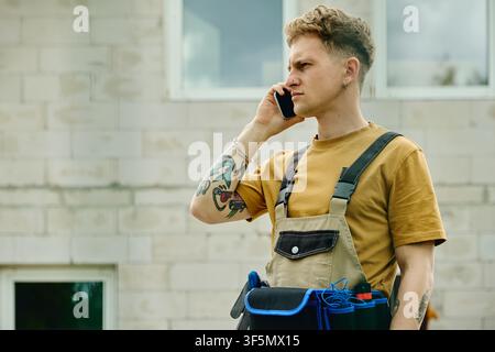 Man in Overalls Talking on Smartphone near House Renovation Site Stock Photo