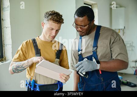 Man and Black Man in Overalls Using Tablet during House Renovation Stock Photo