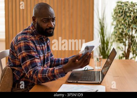 Holding an invoice in one hand and a smartphone in the other, a man carefully reviews his financial documents from home. Stock Photo