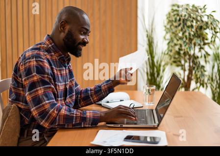 Black man compares paperwork with details on his laptop while working at home. Stock Photo
