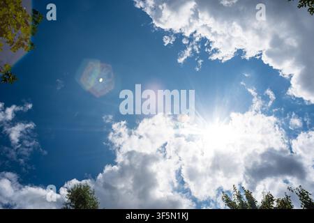 Low angle view looking directly up at the bright midday sun bursting through soft white clouds in a vibrant blue sky framed by tree leaves at the edge Stock Photo