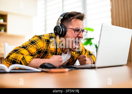 A frustrated young man wearing headphones leans toward his laptop with clenched fists, reacting intensely to what he sees on the screen. Stock Photo