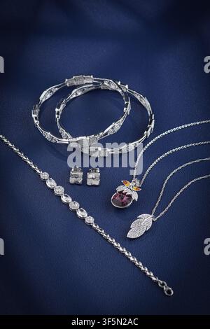 Luxury sterling silver jewelry set featuring bracelets, necklaces, earrings and gemstone pendant displayed on elegant dark blue fabric background Stock Photo