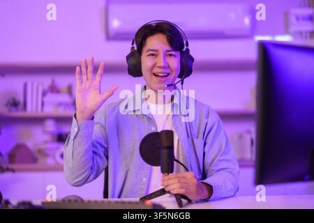 Young Asian man wearing headphones waving at camera during live stream for content creation Stock Photo