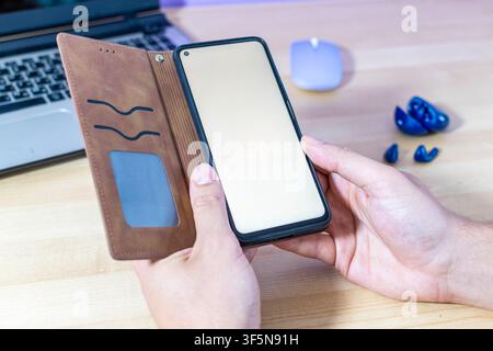 A person holds a smartphone in a brown case at their desk. Blue wireless earbuds and a laptop are in the background. Blank screen mockup. Stock Photo