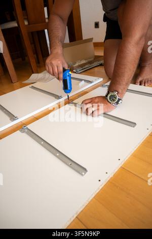 Man assembling flat-pack furniture at home, using an electric screwdriver to attach drawer slides Stock Photo