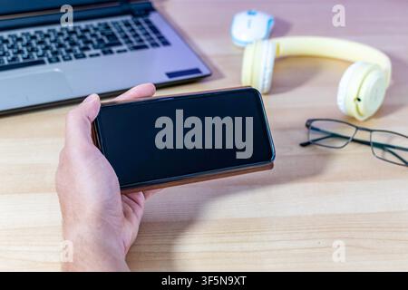 A man holds a mobile phone in a brown case in landscape mode over his desk. The black screen is ideal for mockups. Stock Photo