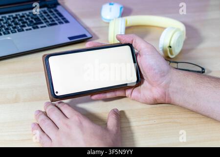 A person holds a mobile phone in a brown wallet case horizontally. The blank screen provides mockup space. Desk with a laptop. Stock Photo