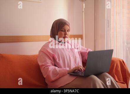 Woman in hijab works on laptop during IV therapy Stock Photo