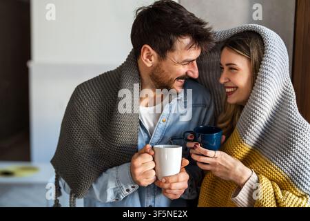 Cheerful young couple covered in blanket embracing while holding cups of tea at home Stock Photo