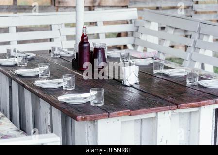 Rustic wooden outdoor dining table set with plates, glasses, cutlery, and bottles of homemade berry juice. Cozy countryside terrace prepared for family lunch, picnic, or gathering in a natural rural setting. Stock Photo