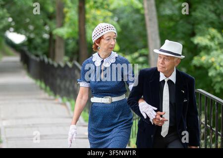 A couple walks hand in hand along a tree-lined path, dressed in elegant vintage outfits. The warm sun filters through the leaves, creating a serene at Stock Photo
