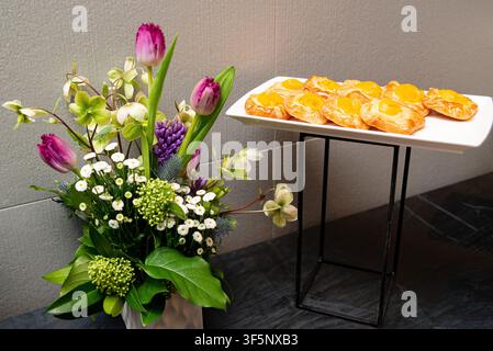 Tulips and white flowers bloom beside golden pastries on a tray. Soft lighting highlights fresh blooms and baked treats. Ideal for cafe, brunch, or ce Stock Photo