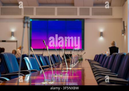 Modern conference room with long table and microphones. Blue chairs line both sides of polished wood table. Large screen glows purple-pink behind pres Stock Photo