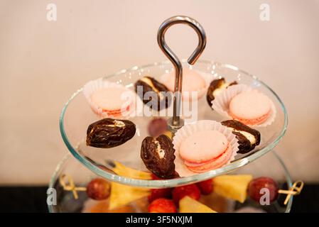 Delicate pink macarons and chocolate-covered dates rest on a glass stand. Fresh fruit skewers add color beneath. Perfect for elegant gatherings or des Stock Photo