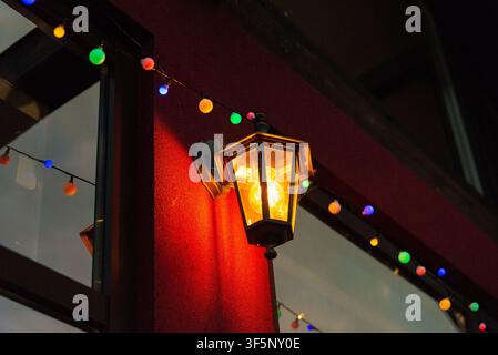 A warmly glowing outdoor wall lantern is mounted on a red exterior wall, illuminating the area with soft yellow light. Festive strings of multicolored Stock Photo