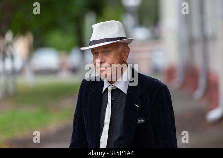 An older man dressed in a dark suit and white hat strolls along a sidewalk, showcasing a unique sense of style amidst a blurred urban backdrop. His ex Stock Photo