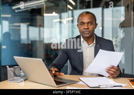 Mature businessman working at a desk in a contemporary office, holding papers and using a laptop, representing professionalism, expertise, and corporate success Stock Photo