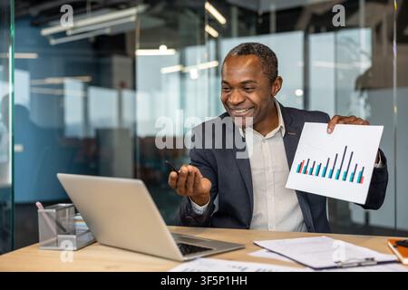 Confident businessman engaging in a virtual meeting, presenting business data and financial growth on a chart document while communicating remotely from a modern office workspace Stock Photo