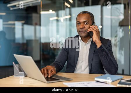 Professional man working at a modern office desk, using his laptop and engaging in a phone call, symbolizing communication, business networking, and efficient work Stock Photo