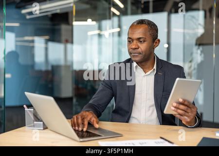 Mature businessman managing tasks, using a laptop and a digital tablet, demonstrating multitasking and efficiency in a contemporary corporate office environment Stock Photo
