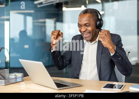 Mature adult man with headphones celebrating achievement in the office, cheering while looking at his laptop screen and expressing excitement about his online success Stock Photo