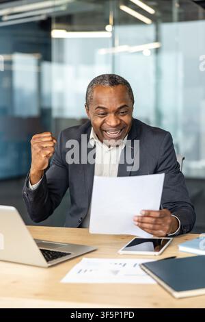 African american businessman celebrating success and achievement, reading a document with an excited expression and clenched fist while sitting at a modern office desk with a laptop and tablet Stock Photo