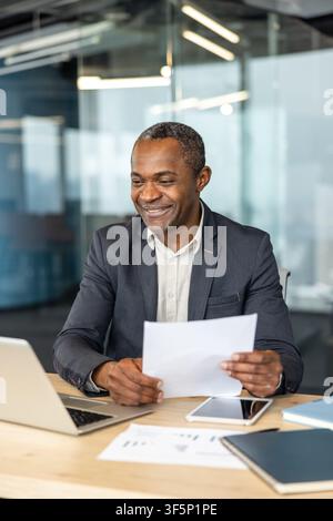 Smiling mature business professional holding and reviewing important documents at a desk with a laptop, tablet, and notebooks, reflecting successful work in a contemporary office setting Stock Photo