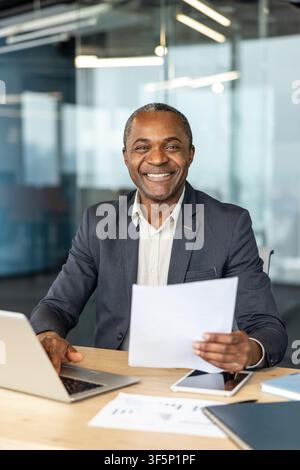 Confident mature businessman working at his desk, happily looking at the camera while holding paperwork and using a laptop in a modern corporate office setting Stock Photo