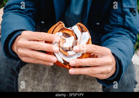 a close-up of a man holding a traditional danish kanelsnegl with both hands, revealing its cinnamon swirls, glossy white icing and a fresh bite taken Stock Photo