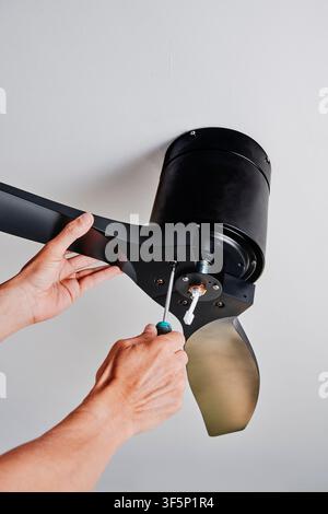 from directly below, a man secures the central housing of a black ceiling fan with a screwdriver as he steadies one curved blade during installation, Stock Photo