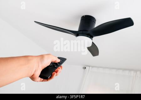 a man uses a remote control to switch on a black three blade ceiling fan with integrated white light, mounted beneath a plain white ceiling in a brigh Stock Photo