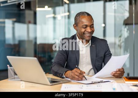 Mature businessman sitting at a desk in a modern office, happily writing on documents and reviewing paperwork, representing concepts of success, professionalism, and administrative work Stock Photo