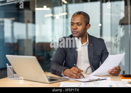 Mature professional businessman sitting at desk in a contemporary office, looking away while reviewing documents and making notes, showcasing focus and dedication to his work Stock Photo