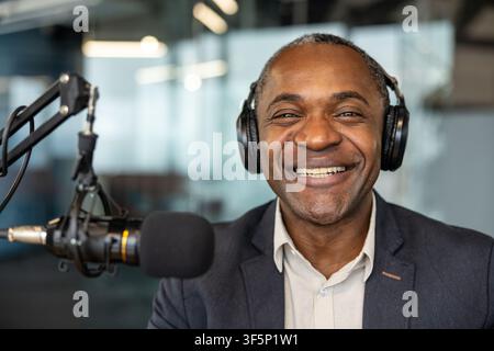 Man wearing headphones and smiling while speaking into a microphone, recording or streaming a podcast or radio show in a professional studio Stock Photo