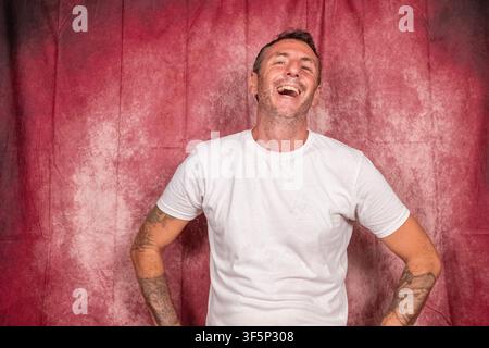 A man laughs cheerfully, the man has short hair, a short beard, wears a white T-shirt, has tattooed arms and is portrayed on a red background Stock Photo