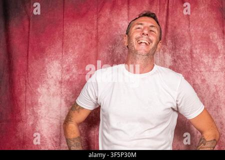 A man laughs cheerfully, the man has short hair, a short beard, wears a white T-shirt, has tattooed arms and is portrayed on a red background Stock Photo