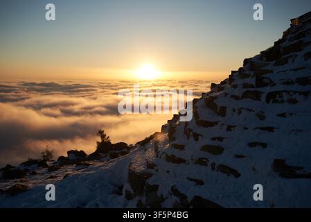 Sunrise over snow-covered mountain peak. Warm golden light of rising sun bathes clouds in soft hues of orange, pink. Sky transitions from deep blue to gentle pastel. Stock Photo