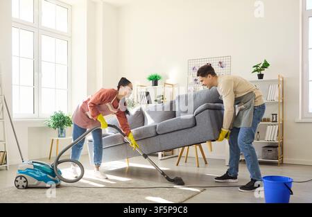 Couple vacuuming under sofa while lifting couch in bright modern living room Stock Photo