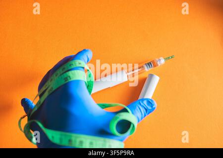 Hand wearing blue glove holding a glp-1 injection pen and green measuring tape on orange background Stock Photo