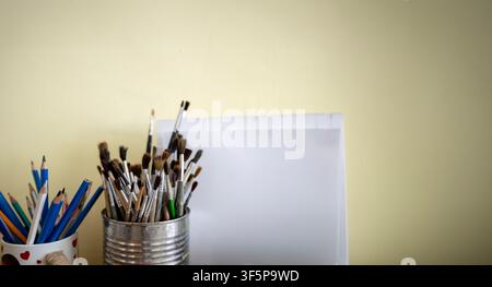Artistic tools displayed in metal containers next to a blank paper surface against a neutral yellow wall various brushes and wooden pencils for creati Stock Photo