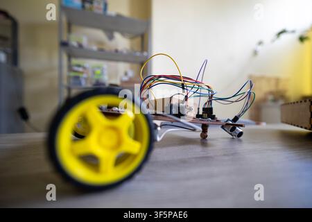 Robotic vehicle prototype with yellow wheels and colorful wires sitting on a wooden desk inside a workshop area storage shelves in the background whil Stock Photo