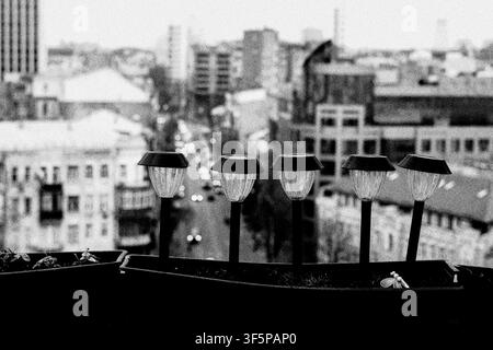 Outdoor solar garden lights lined up in a planter on a terrace balcony overlooking a blurry urban cityscape with blurred moving vehicles and distant b Stock Photo