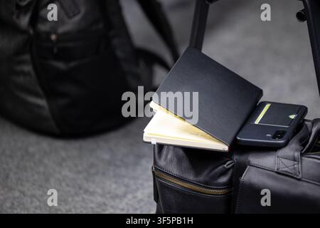 Close up a leather bag holding an open notebook and a mobile device with a card slot while resting on a hard grey floor near another black pack in pro Stock Photo