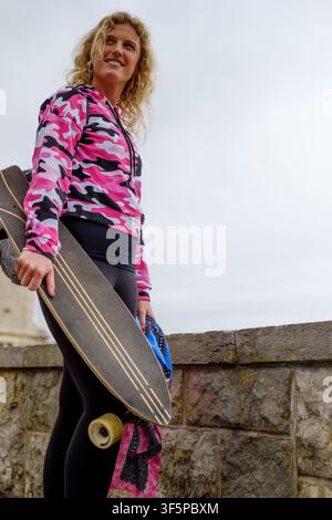 Young woman standing with longboard, smiling, dressed in pink camouflage hoodie Stock Photo