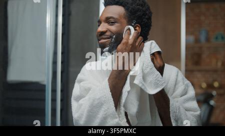 Mirror reflection of happy positive smiling African American man wiping cheeks after shaving shave gel foam soap optimistic male guy wipe foamy beard Stock Photo