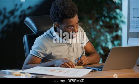 Concentrated African American business man architect analyzing construction blueprint writing notes with laptop at night office focused male guy Stock Photo